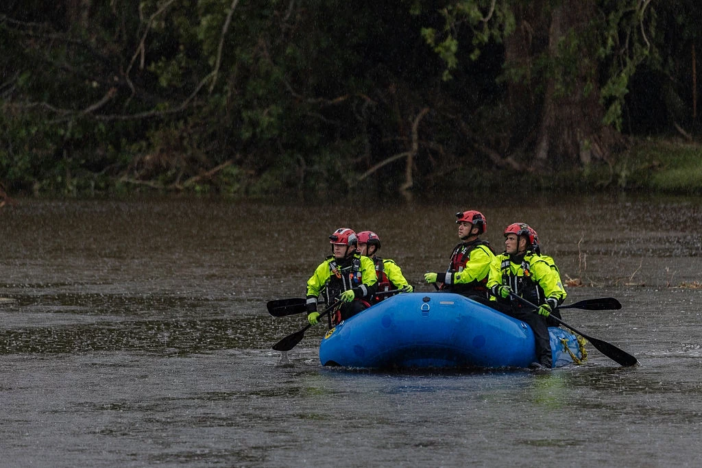 Poszukiwania zaginionych w Camp Mystic w hrabstwie Kerr Czterech ratowników w żółtych kurtkach i kaskach siedzi w niebieskim pontonie płynącym po zalanej rzece, wokół widoczna jest bujna roślinność.