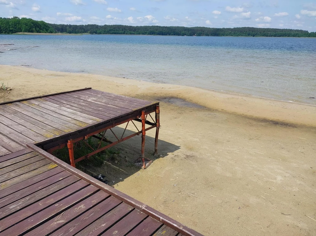 Jezioro Świętajno Narckie Drewniany pomost częściowo nad suchym piaskiem, brak wody pod jego częścią, spokojna linia brzegowa jeziora, daleko widoczny las po drugiej stronie jeziora, niebo z drobnymi chmurami.