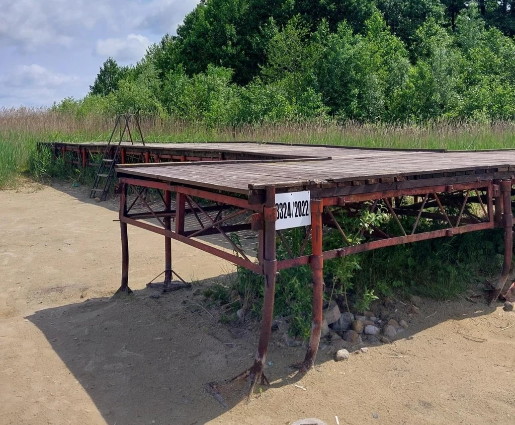 Konstrukcja sceniczna wykonana z metalowych i drewnianych elementów ustawiona na zewnątrz na ziemi, otoczona zielenią i trawą, z widocznym numerem identyfikacyjnym na jednym z elementów stelaża.