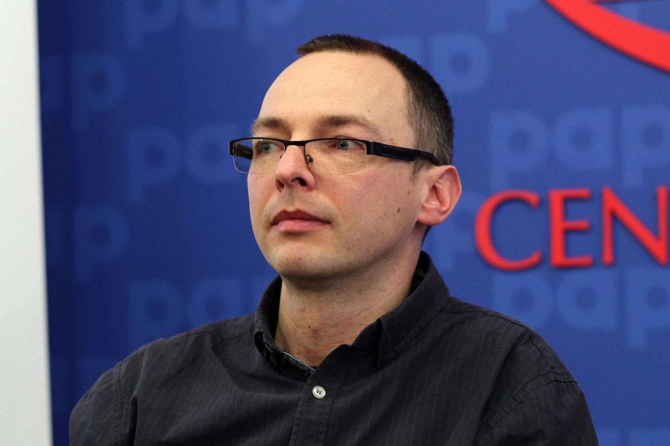 Mężczyzna w okularach z krótkimi ciemnymi włosami, ubrany w ciemną koszulę, siedzi na tle niebieskiej ścianki z napisem i czerwonym elementem graficznym.