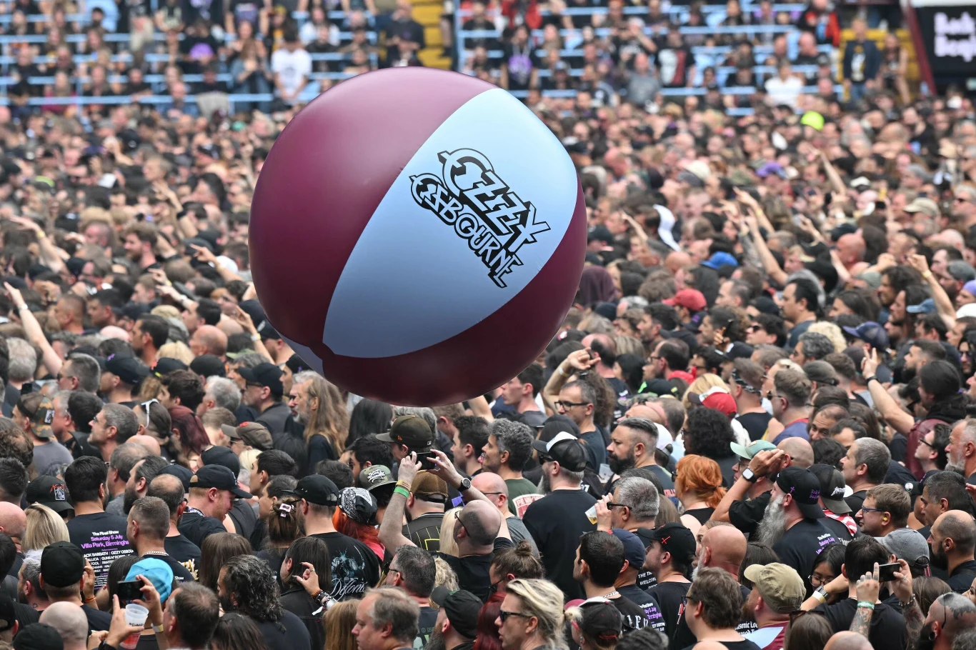 Pożegnanie Black Sabbath odbyło się w Birmingham Tłum ludzi na koncercie, nad którym unosi się duża plażowa piłka z napisem Ozzy Osbourne, widoczne różne osoby skupione na wydarzeniu, atmosfera zabawy i energii.