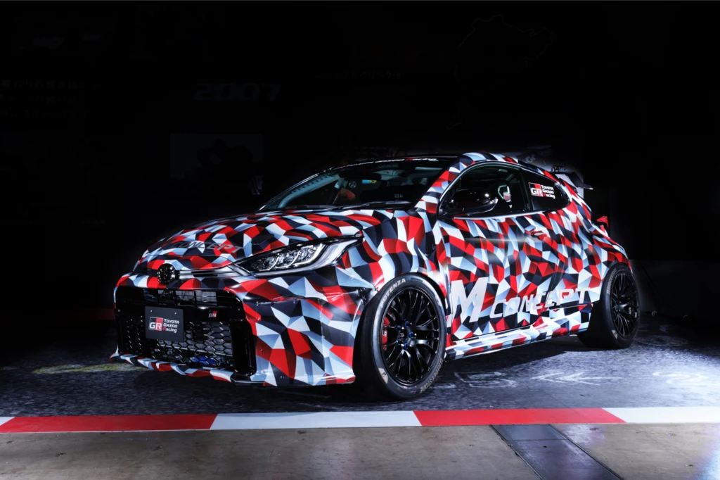 Nowa Toyota Yaris GR M Concept napędzana jest nową jednostką czterocylindrową o mocy około 450 KM. Samochód sportowy z agresywnym body kitem, pokryty wielokolorowym, geometrycznym kamuflażem, stojący na torze, podkreślony efektownym oświetleniem, nowoczesne felgi oraz detale wzmacniają wyścigowy charakter pojazdu.