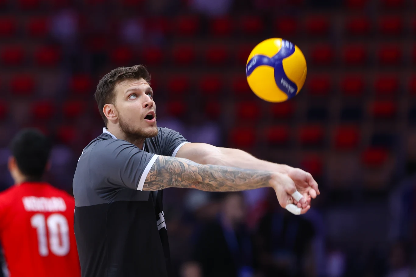 Arthur Szwarc w reprezentacji Kanady Zawodnik odbijający piłkę do siatkówki sposobem dolnym podczas meczu lub treningu, w tle rozmyty zawodnik w czerwonej koszulce z numerem 10 oraz widownia.