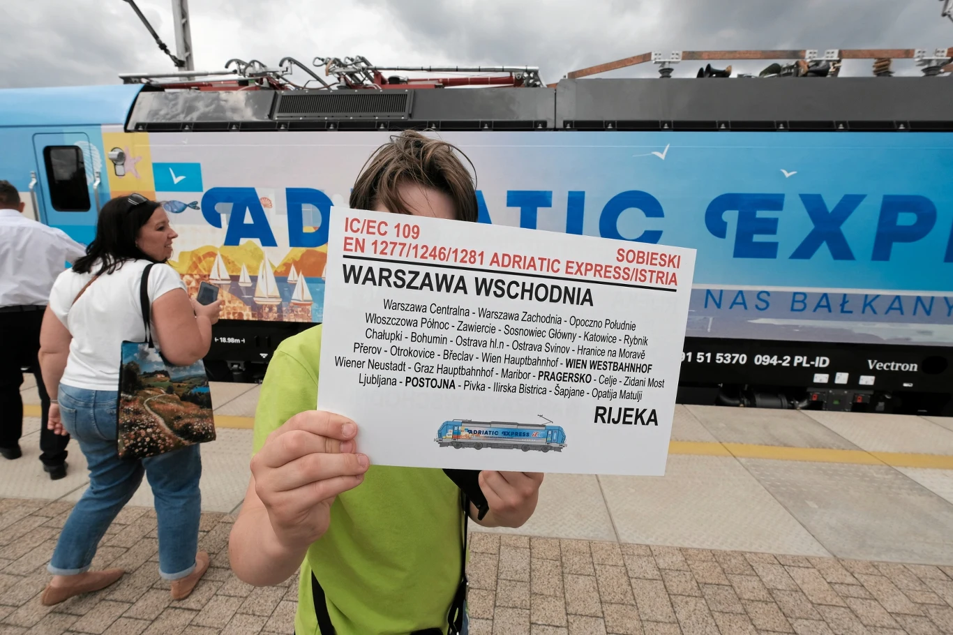 Adriatic Express PKP Intercity wraca do Chorwacji. Kiedy sprzedaż biletów? Osoba w zielonej koszulce trzyma kartkę z rozkładem Adriatic Express na tle niebieskiej lokomotywy i wagonu na peronie.