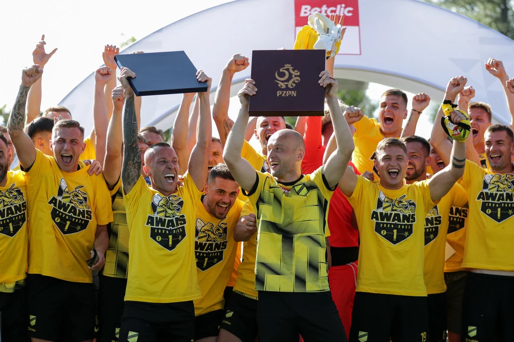 Zawodnicy Wieczystej Kraków świętujący awans do Betclic I ligi Grupa piłkarzy w żółtych koszulkach świętuje awans, unosząc trofeum oraz dyplom Polskiego Związku Piłki Nożnej, wszyscy uśmiechnięci i wyraźnie podekscytowani sukcesem