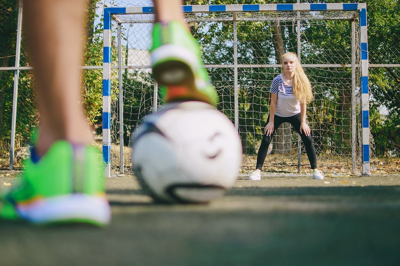 Dziewczynka w pozycji bramkarza stoi na tle bramki do piłki nożnej, skoncentrowana na piłce, którą zaraz kopnie osoba znajdująca się na pierwszym planie. W tle metalowa siatka i drzewa.