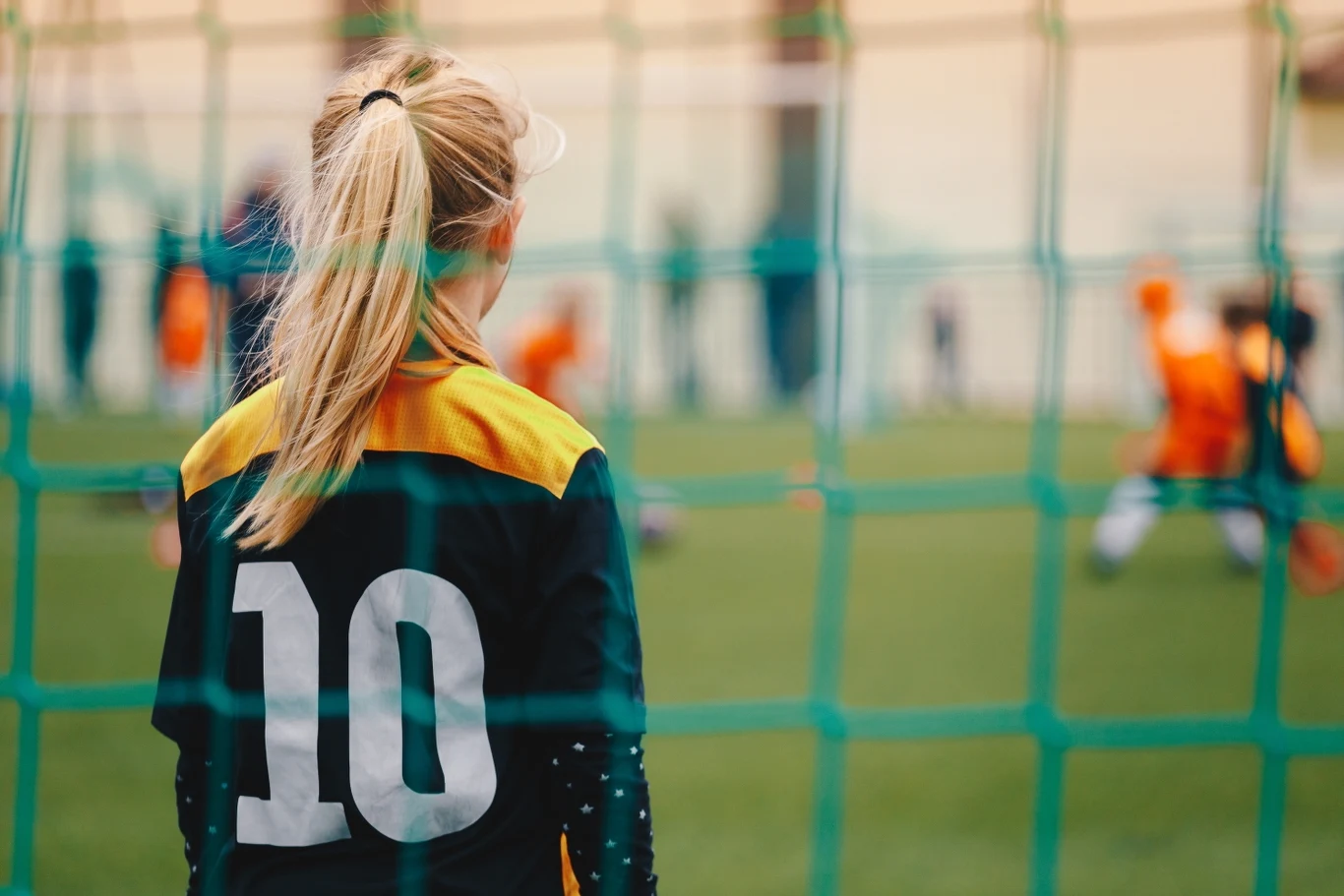 Dziewczynka w koszulce z numerem 10 stoi na boisku piłkarskim w pobliżu bramki, obserwując grę innych dzieci ubranych w pomarańczowe stroje sportowe, całość widoczna przez zieloną siatkę bramkarską.