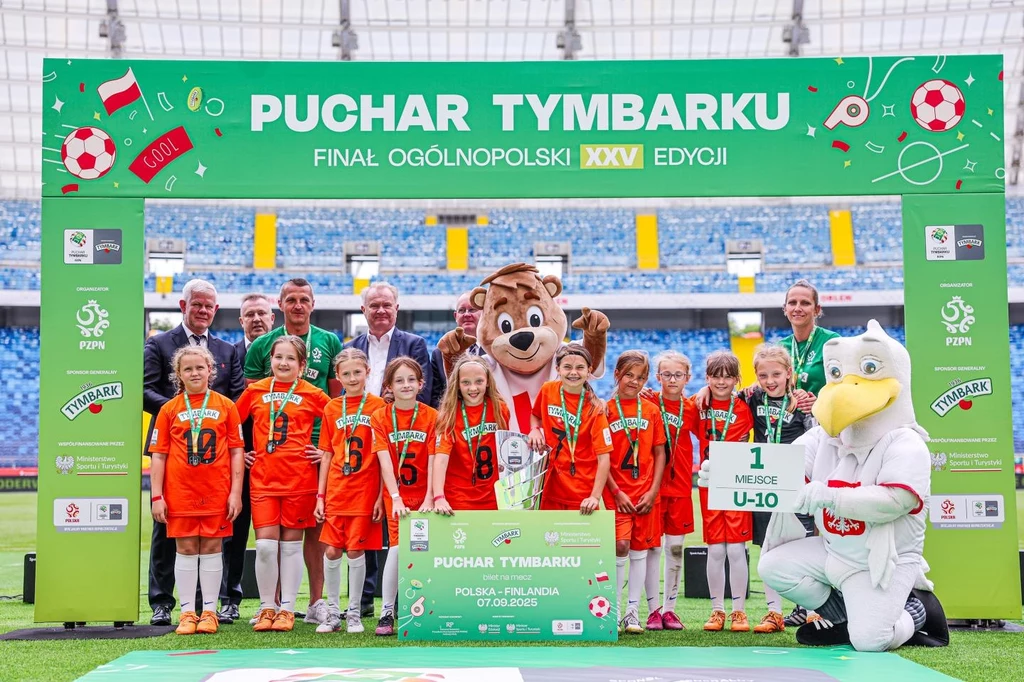 Finał XXV edycji Pucharu Tymbarku: fot. Cyfra Sport Dziewczęca drużyna piłkarska w pomarańczowych strojach świętuje zwycięstwo w finale turnieju, stojąc pod banerem z napisem Puchar Tymbarku, obok maskotek i dorosłych opiekunów, na stadionie piłkarskim.
