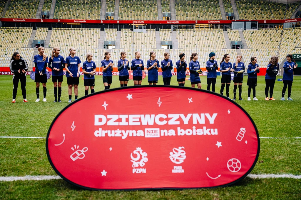 PZPN wspiera dziewczynki i kobiety poprzez takie działania jak program grantowy „Kobiety w grze”: fot. Mateusz Słodkowski / Cyfrasport Dziewczęca drużyna piłkarska ustawiona w szeregu na murawie stadionu sportowego, w tle trybuny, na pierwszym planie duży czerwony baner z napisem promującym udział dziewczyn w piłce nożnej.