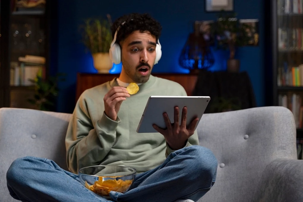 Seriale i filmy bez długoterminowych umów Młody mężczyzna z kręconymi włosami i zarostem siedzi na szarej sofie, ma na sobie jasne słuchawki i trzyma tablet. W jednej ręce trzyma chipsa, na jego twarzy widać zaskoczenie. Przed nim stoi miska pełna przekąsek, a tło jest rozmyte z elementami dom...