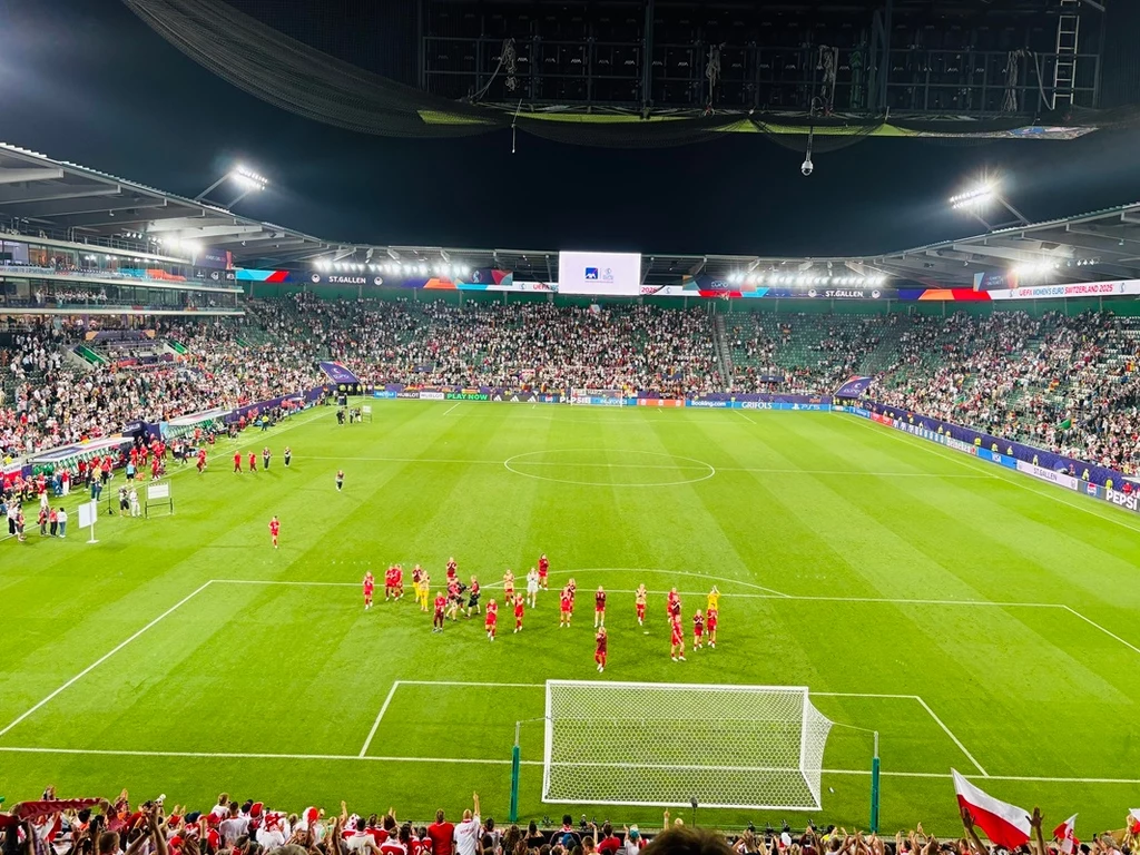 Piłkarze w czerwonych strojach świętujący sukces na murawie stadionu piłkarskiego, trybuny pełne kibiców, stadion oświetlony reflektorami, w tle duży ekran i tłum entuzjastycznych widzów.