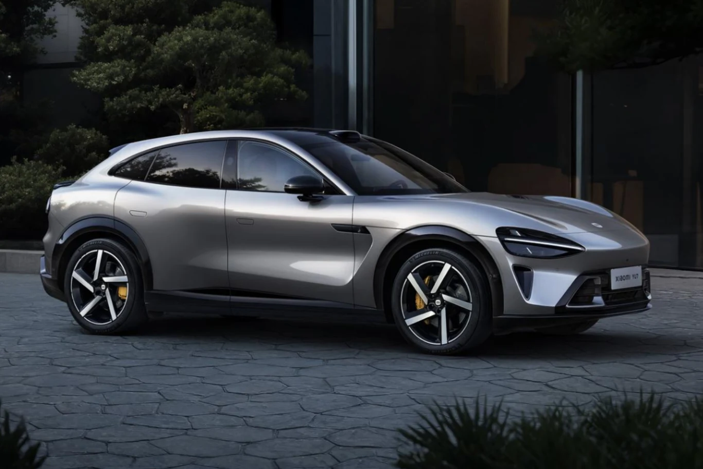 Nowoczesny srebrny samochód elektryczny o sportowej sylwetce zaparkowany na tle budynku z dużymi szybami i zielenią w tle.