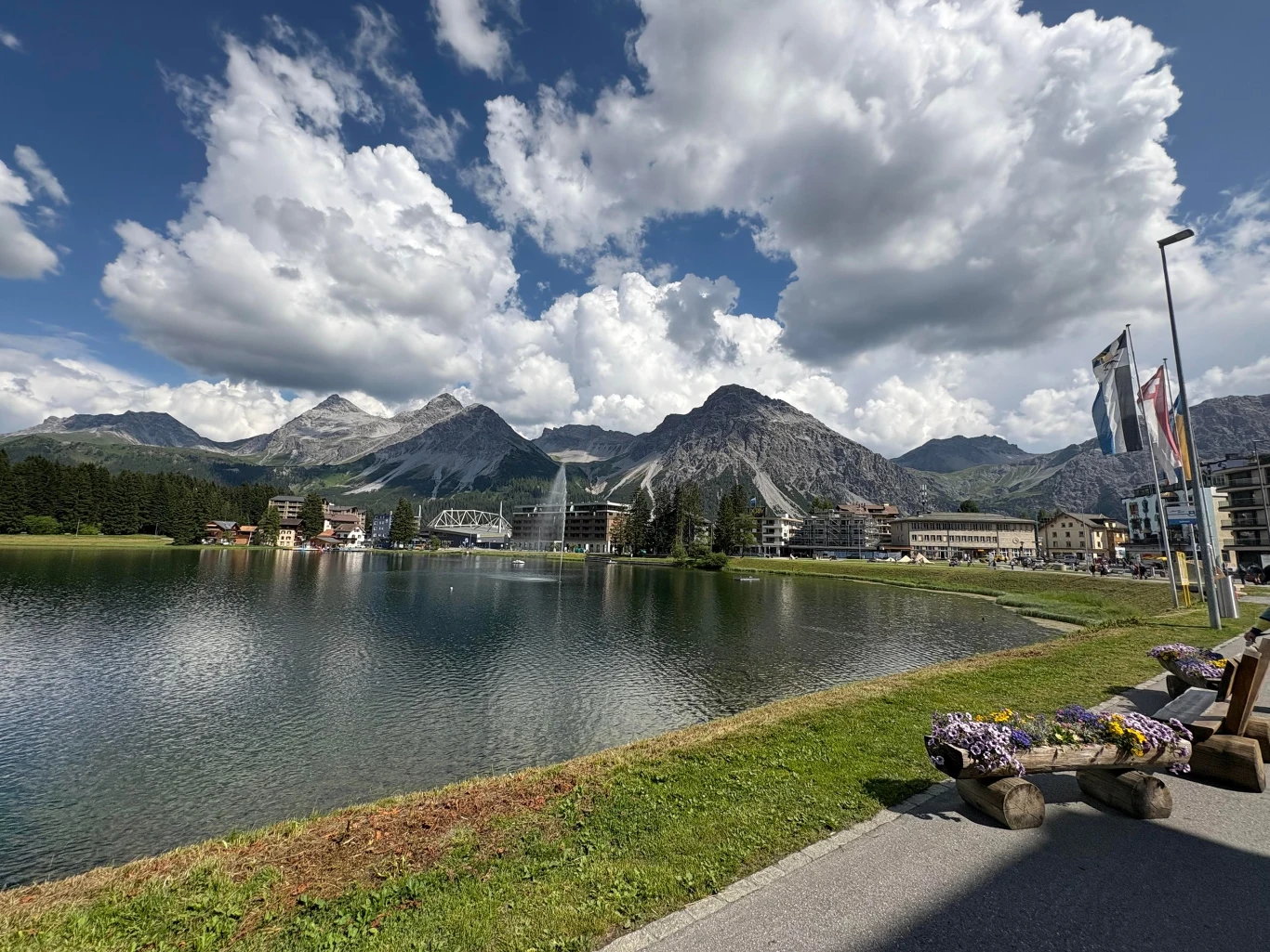 Arosa to popularny górski kurort Malownicze jezioro otoczone górskimi szczytami, nowoczesna zabudowa miejska w tle, po prawej stronie ławki z kwiatami oraz flagi na masztach, widoczne dynamiczne chmury na niebie.