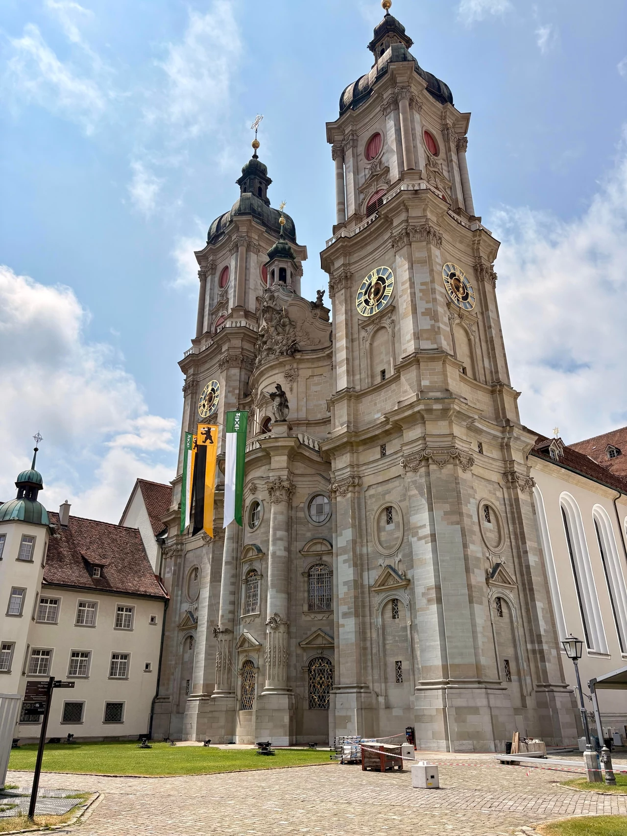 Monumentalna barokowa katedra o dwóch wysokich wieżach z zegarami, przestrzenne wejście otoczone flagami, plac wyłożony brukiem, fragmenty przylegających budynków oraz błękitne niebo z lekkimi chmurami.