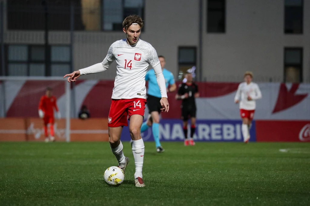 Kacper Potulski Piłkarz w białej koszulce z numerem 14 i czerwonych spodenkach prowadzi piłkę na boisku, na tle innych zawodników oraz budynków, w tle reklamy i bramka.