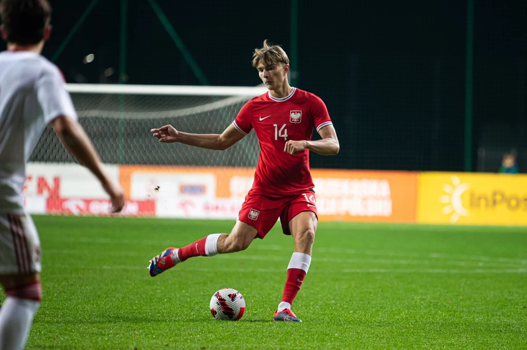 Kacper Potulski Piłkarz w czerwonym stroju reprezentacji Polski wykonuje podanie podczas meczu piłkarskiego na zielonej murawie stadionu, w tle widoczna bramka oraz fragment innego zawodnika.
