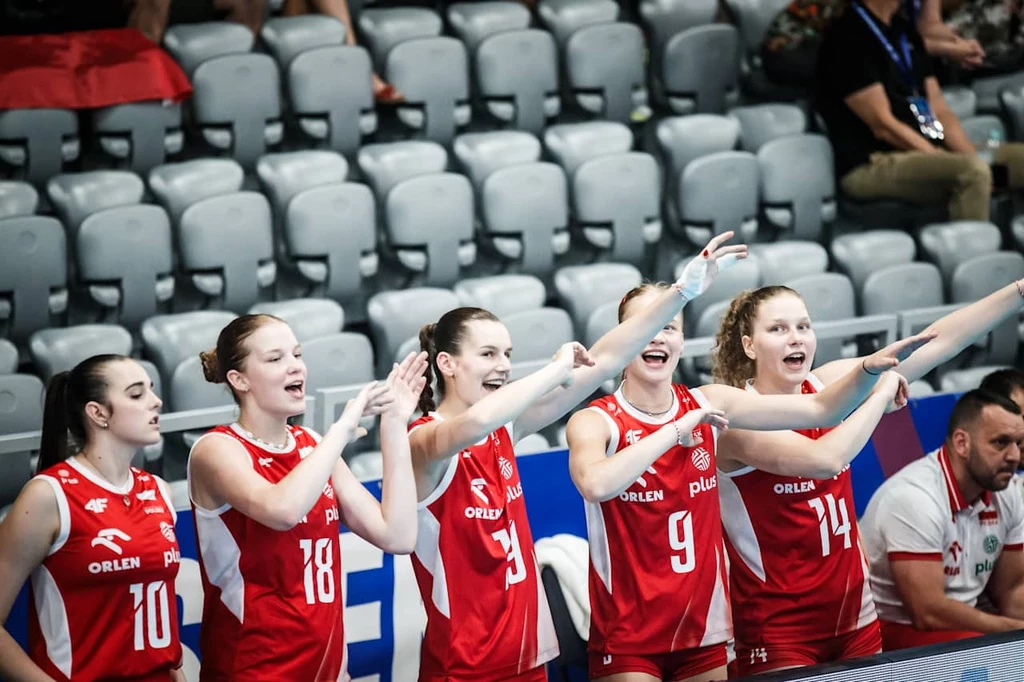 Grupa zawodniczek drużyny siatkarskiej w czerwonych strojach dopingujących swoją drużynę na trybunach, wszystkie uśmiechnięte i zaangażowane, w tle puste rzędy krzeseł oraz trener obserwujący wydarzenia.