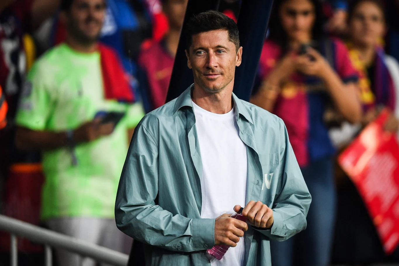 Mężczyzna w jasnej koszulce i rozpiętej koszuli stoi wśród kibiców na stadionie, w tle zarysowane osoby oraz barwne flagi i koszulki sportowe.