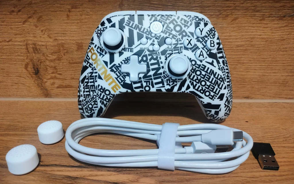 PowerA OPS V1 Fortnite Edition Bezprzewodowy kontroler do gier z napisem 'Fortnite' i dynamicznym wzorem czarno-białych napisów, w zestawie z kablem USB, końcówkami na drążki oraz adapterem USB, całość na tle drewnianej powierzchni.