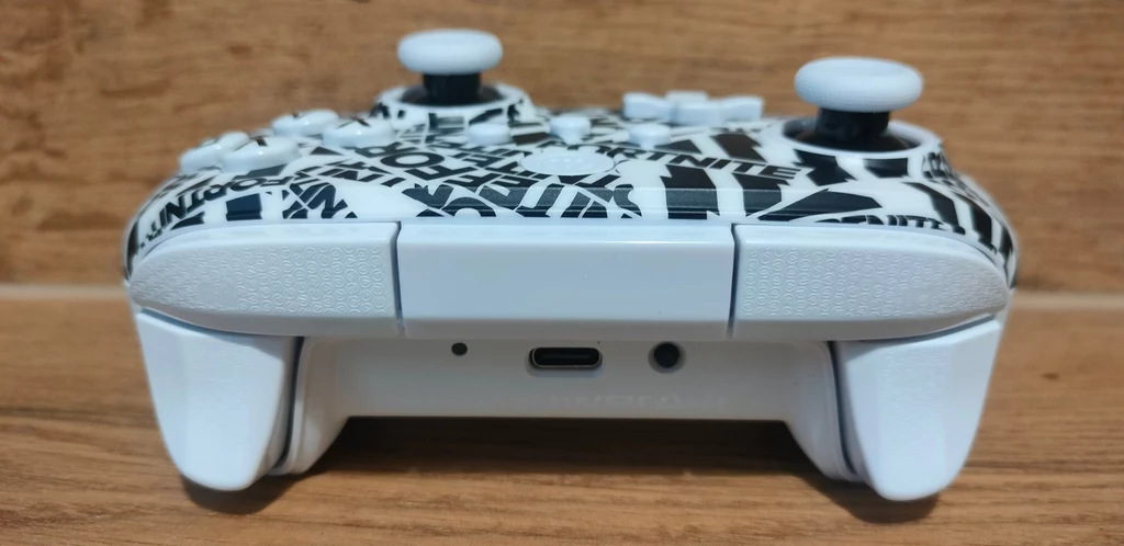 PowerA OPS V1 Fortnite Edition Biały kontroler do gier z czarno-białym wzorem na przodzie, leżący na drewnianej powierzchni, z widocznym portem ładowania USB-C pośrodku dolnej części obudowy.