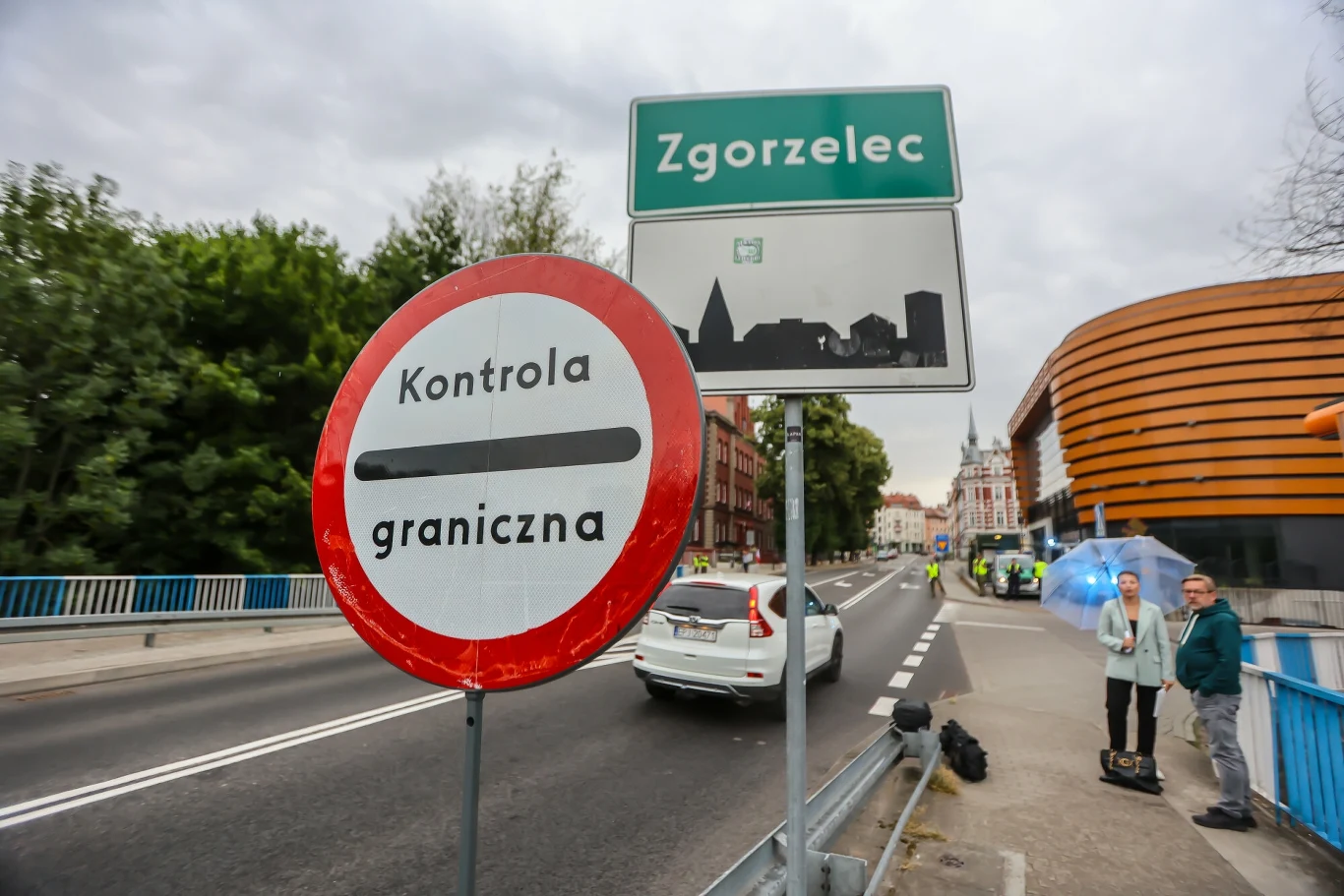 Władze Zgorzelca zapowiadają skargę do KE na kontrole graniczne po niemieckiej stronie Znak kontroli granicznej oraz tablica z nazwą miejscowości Zgorzelec ustawione przy drodze na moście granicznym, po lewej stronie widoczna roślinność, po prawej nowoczesny budynek, w tle samochód i kilka osób na chodniku.