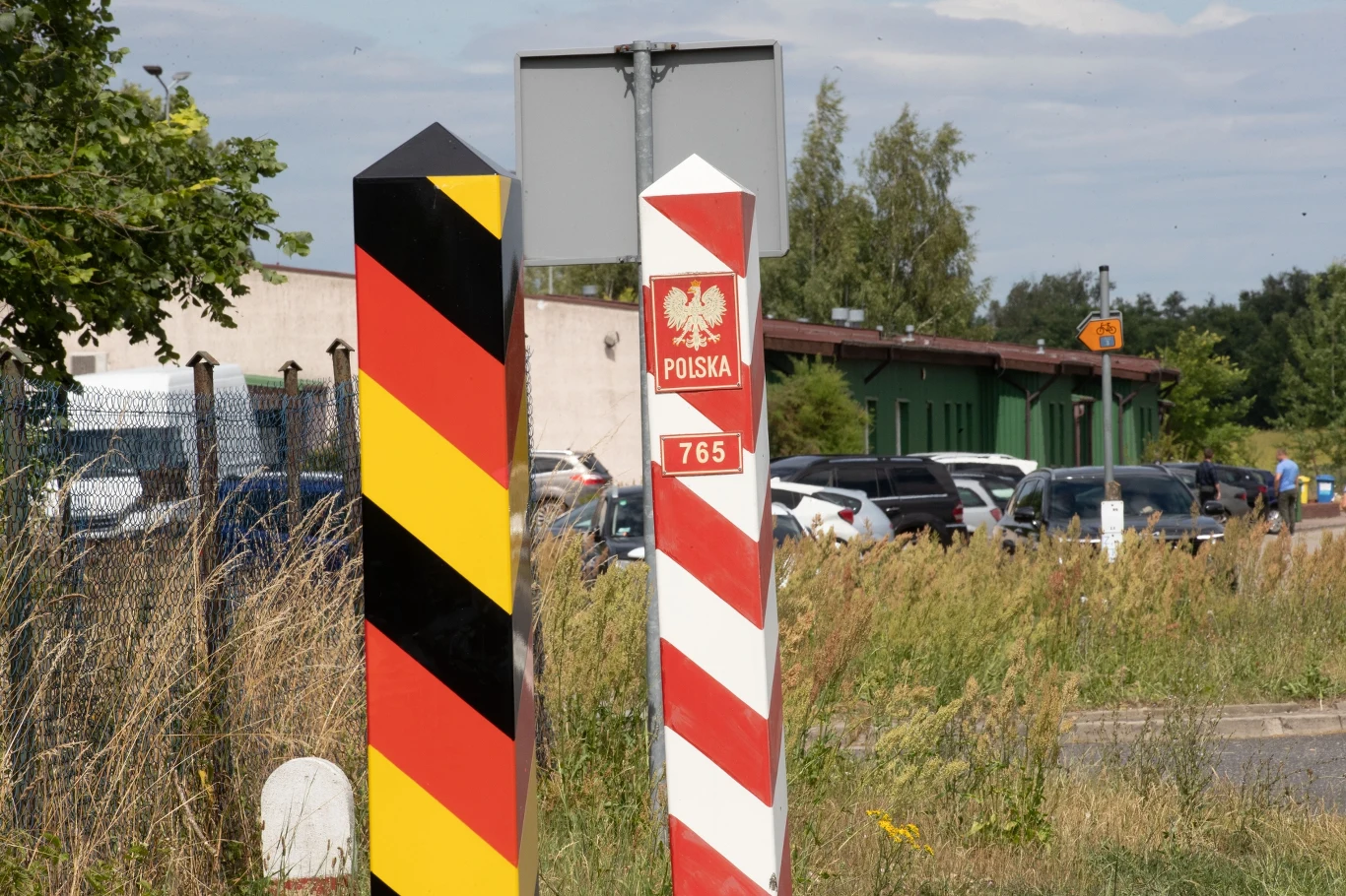 Polsko-niemieckie przejście graniczne Rosówek - Rosow Słupki graniczne Polski i Niemiec stojące obok siebie na tle budynków, zaparkowanych samochodów i roślinności, symbolizujące przejście graniczne między dwoma państwami.