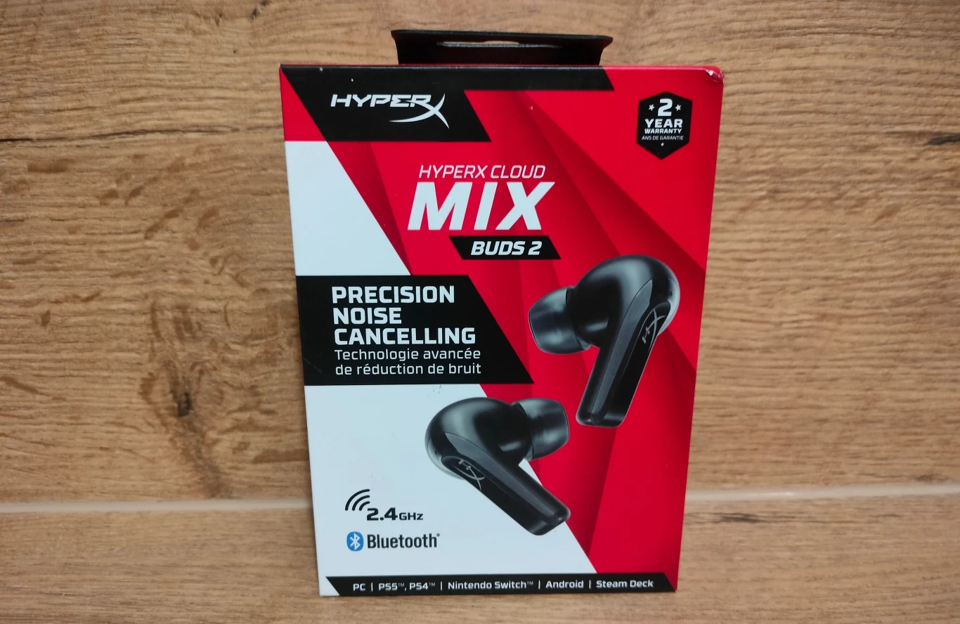 HyperX Cloud Mix Buds 2 Opakowanie słuchawek bezprzewodowych HyperX Cloud Mix Buds 2 w czarno-czerwonej kolorystyce, z czytelnymi informacjami o technologii redukcji hałasu i obsłudze Bluetooth, na tle drewnianej powierzchni.