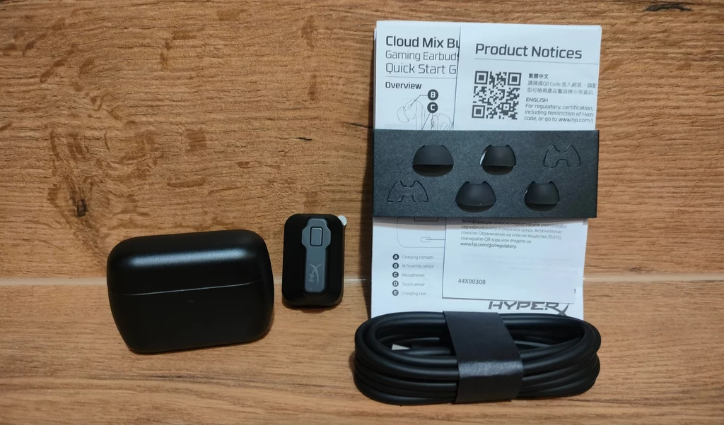 HyperX Cloud Mix Buds 2 Czarna etui na słuchawki, moduł sterujący, silikonowe nakładki, czarny kabel oraz instrukcja obsługi do słuchawek HyperX ułożone na drewnianej powierzchni.