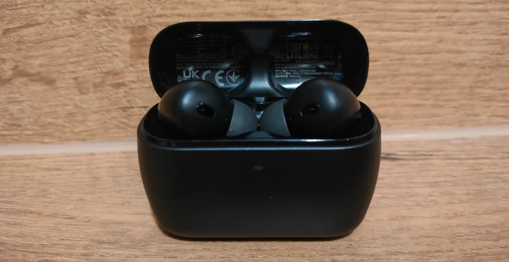 HyperX Cloud Mix Buds 2 Czarne bezprzewodowe słuchawki douszne umieszczone w otwartym etui ładującym na tle drewnianej powierzchni.