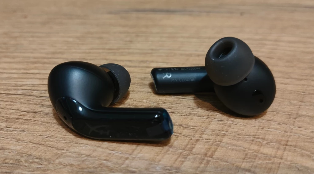HyperX Cloud Mix Buds 2 Dwie bezprzewodowe słuchawki douszne w czarnym kolorze leżące na drewnianej powierzchni, dobrze widoczne detale silikonowych końcówek oraz powierzchni obudowy.