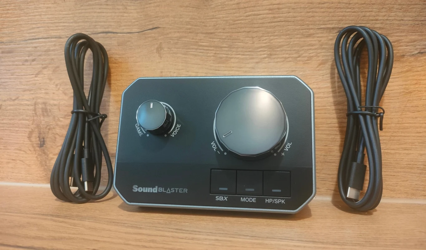 Creative Sound Blaster G8 Panel sterowania audio Sound Blaster z dużym i małym pokrętłem oraz trzema przyciskami funkcyjnymi, po bokach ułożone dwa kable zakończone wtyczkami USB, całość na tle drewnianej powierzchni.