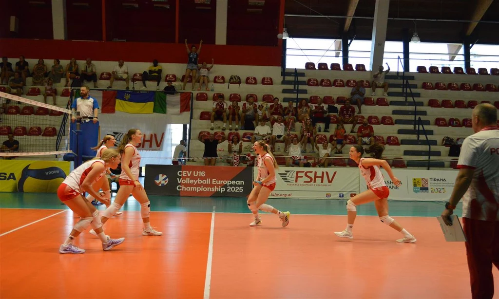 Zawodniczki drużyny siatkarskiej w sportowych strojach podczas meczu na hali, w tle sędzia na podwyższeniu, kibice na trybunach oraz baner informujący o Mistrzostwach Europy w siatkówce 2025.