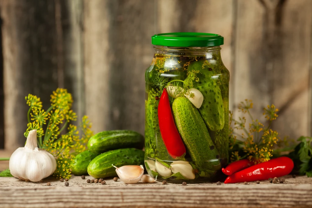 Dodatek chili urozmaici tradycyjne ogórki kiszone Słoik z kiszonymi ogórkami wypełniony także papryczkami chili, ząbkami czosnku i koperkiem, ustawiony na drewnianym stole w otoczeniu świeżych ogórków, czosnku, papryczek i przypraw.