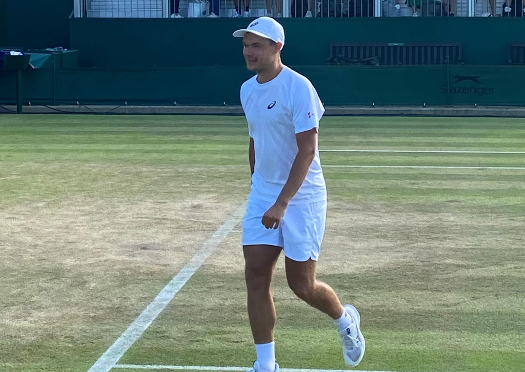 Kamil Majchrzak Mężczyzna w sportowym stroju tenisowym na korcie trawiastym, uśmiechnięty, z czapką na głowie i rakietą tenisową w ręce, w tle widoczna trybuna stadionowa.