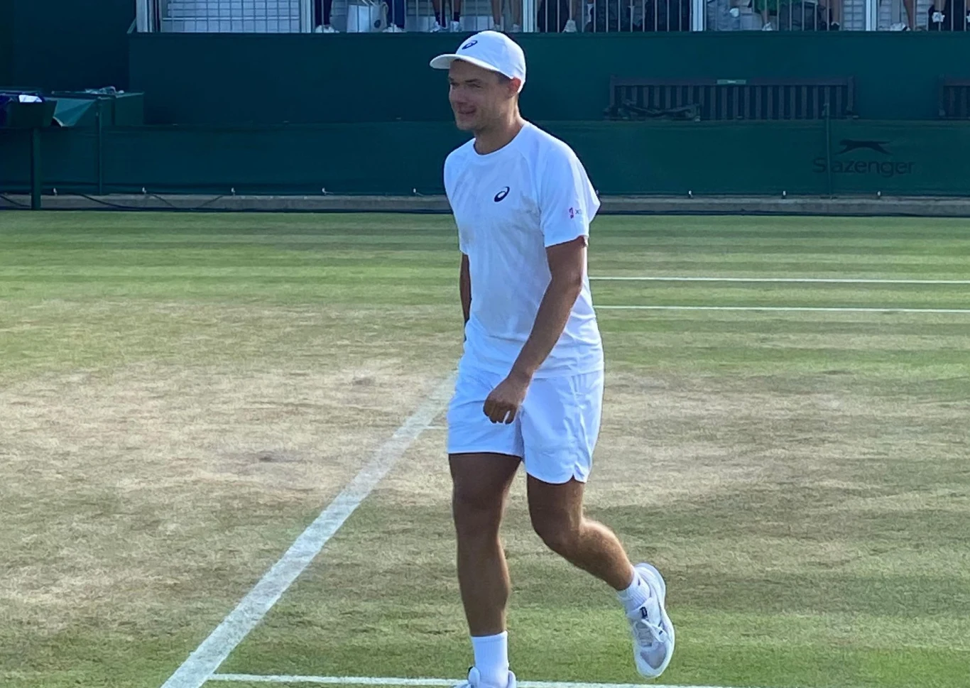 Mężczyzna w sportowym stroju tenisowym na korcie trawiastym, uśmiechnięty, z czapką na głowie i rakietą tenisową w ręce, w tle widoczna trybuna stadionowa.