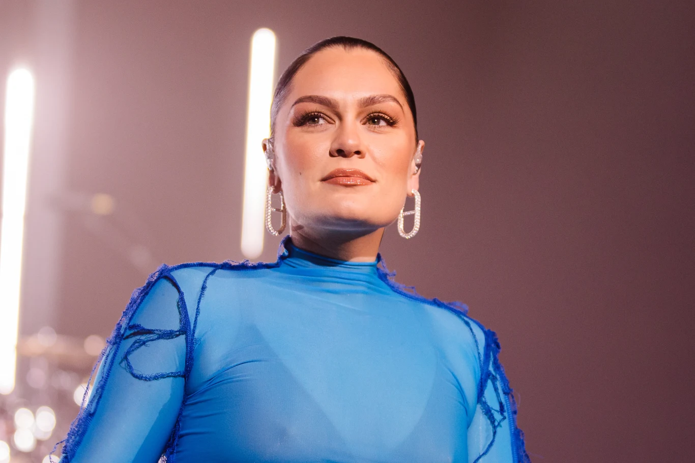 Jessie J przekazała nowe wieści o swoim zdrowiu Kobieta w niebieskiej, przezroczystej bluzce z dużymi kolczykami stoi na tle rozmytego światła jako element sceny koncertowej, spokojny wyraz twarzy, włosy zaczesane do tyłu.