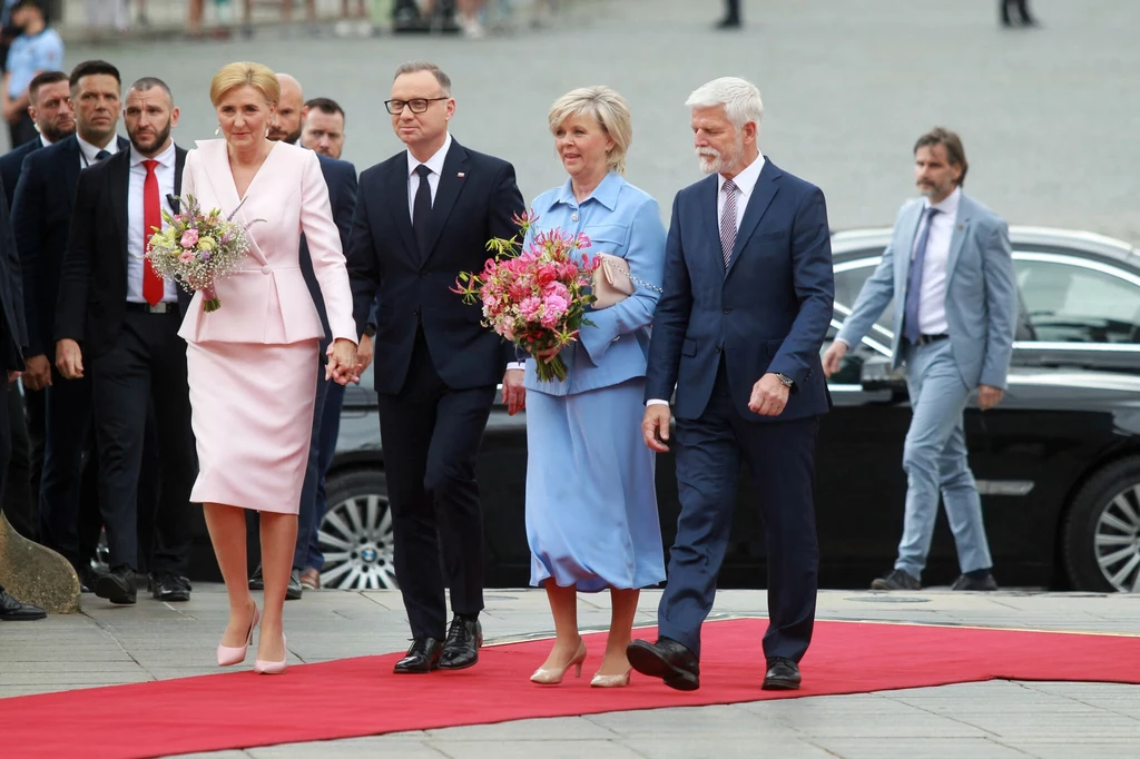 Wizyta polskiej pary prezydenckiej w Pradze, 2025 r. Cztery osoby ubrane elegancko idą po czerwonym dywanie, dwie kobiety trzymają w rękach bukiety kwiatów, w tle widoczni są ochroniarze oraz samochód służbowy.