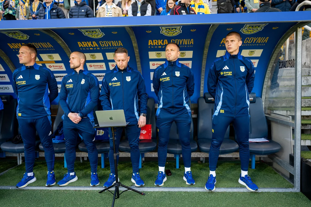 Sztab szkoleniowy Arki Gdynia Pięciu mężczyzn ubranych w klubowe dresy Arki Gdynia stoi pod ławką rezerwowych na stadionie piłkarskim, przy ich nogach stoi laptop, w tle widoczni kibice i część trybun.