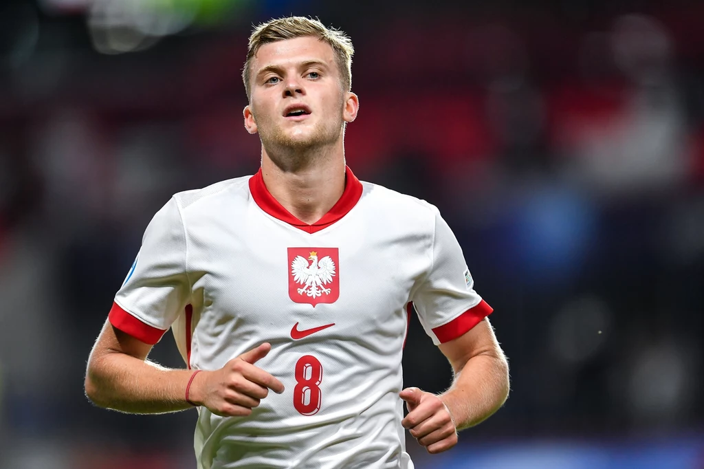 Młody piłkarz w białej koszulce reprezentacji Polski z numerem 8 biegnie po boisku, skoncentrowany patrzy przed siebie, w tle niewyraźne trybuny stadionu.
