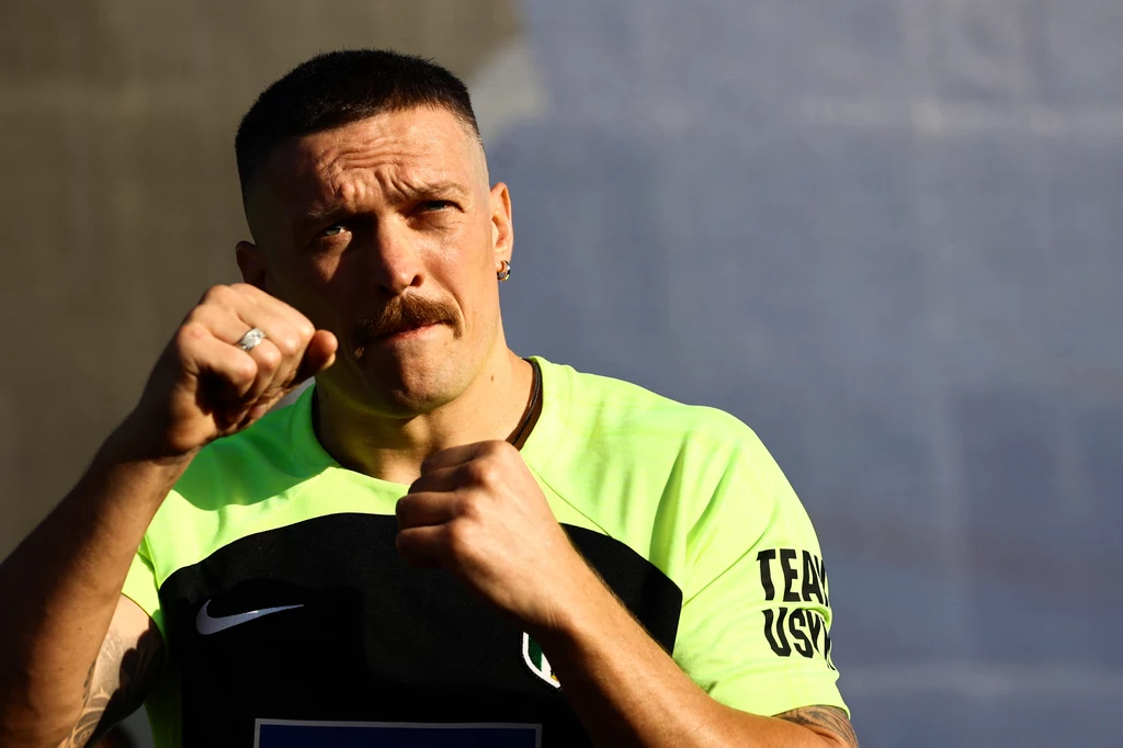 Ołeksandr Usyk Mężczyzna w sportowej koszulce z krótkimi rękawami przyjmuje pozycję bokserską z uniesionymi pięściami, wyraz twarzy skupiony i zacięty, w tle neutralne rozmyte tło.