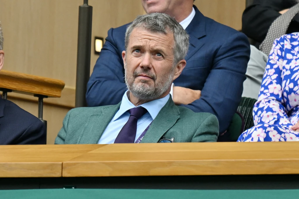 Król Danii Fryderyk X na meczu Igi Świątek i Clary Tauson Mężczyzna w średnim wieku z siwą brodą i krótkimi włosami siedzi w loży widowni w eleganckim zielonym garniturze i fioletowym krawacie, obok innych osób, w tle wnętrze sali.