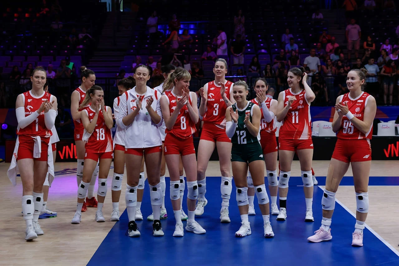 Grupa siatkarek w czerwono-białych strojach drużynowych stoi razem na hali sportowej, uśmiechając się i bijąc brawo po zakończonym meczu, na tle widowni i oświetlonego parkietu.