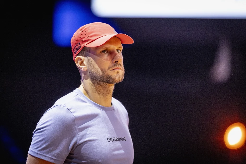 Maciej Ryszczuk Mężczyzna w czerwonej czapce z daszkiem ubrany w jasnofioletową koszulkę sportową z napisem ON RUNNING, skoncentrowany i patrzący w dal, ciemne tło rozświetlone pojedynczym punktem światła.