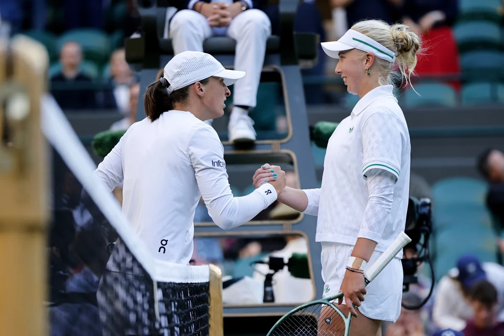 Iga Świątek i Clara Tauson Dwie tenisistki podają sobie dłonie na korcie po zakończonym meczu, jedna ubrana w białą koszulkę i spódniczkę, druga w sportowy strój z długim rękawem, w tle sędzia siedzący na podwyższeniu.