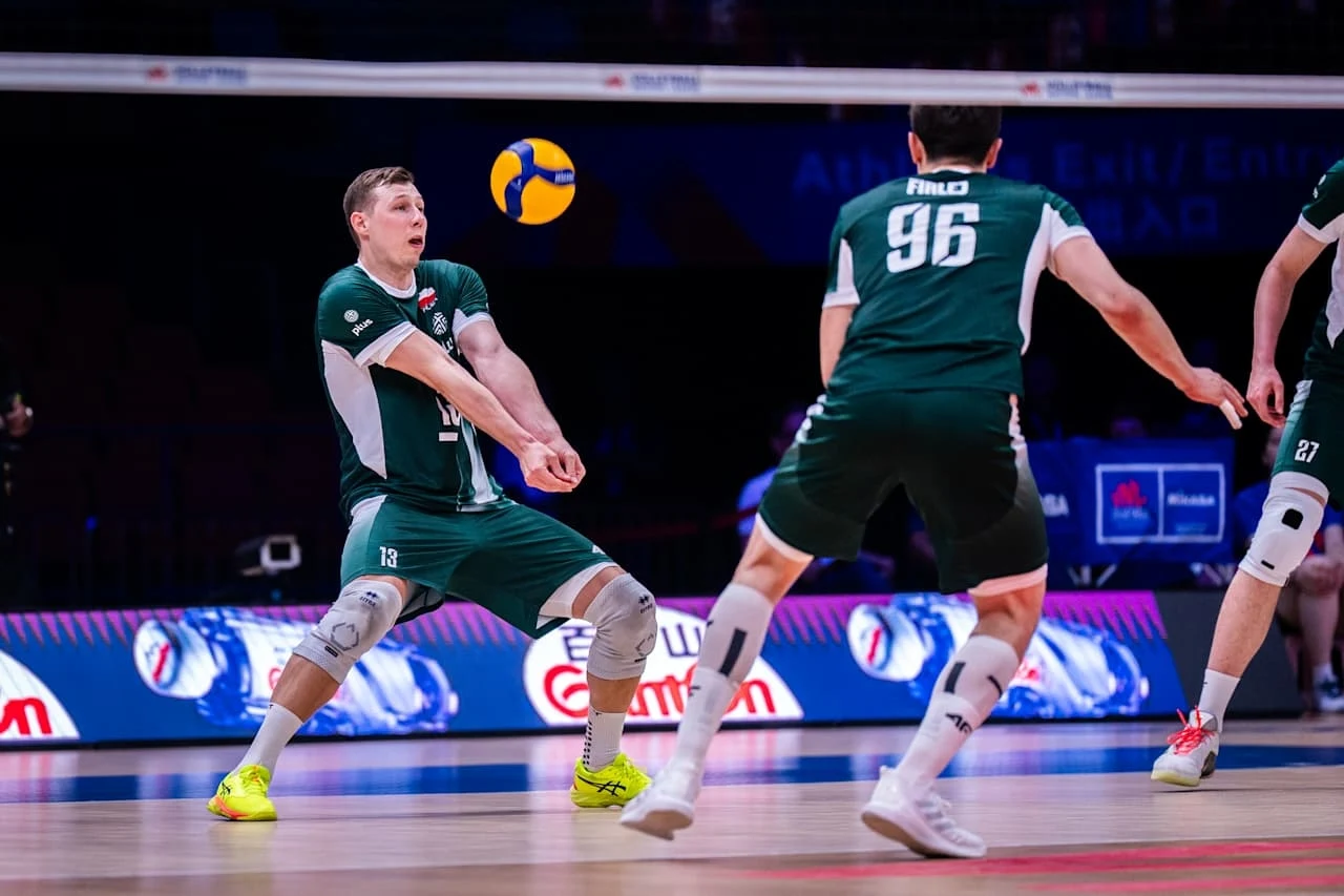 Dwóch zawodników drużyny siatkarskiej w zielonych strojach wykonuje akcję obronną na hali sportowej, skupiając się na odbiciu lecącej piłki.