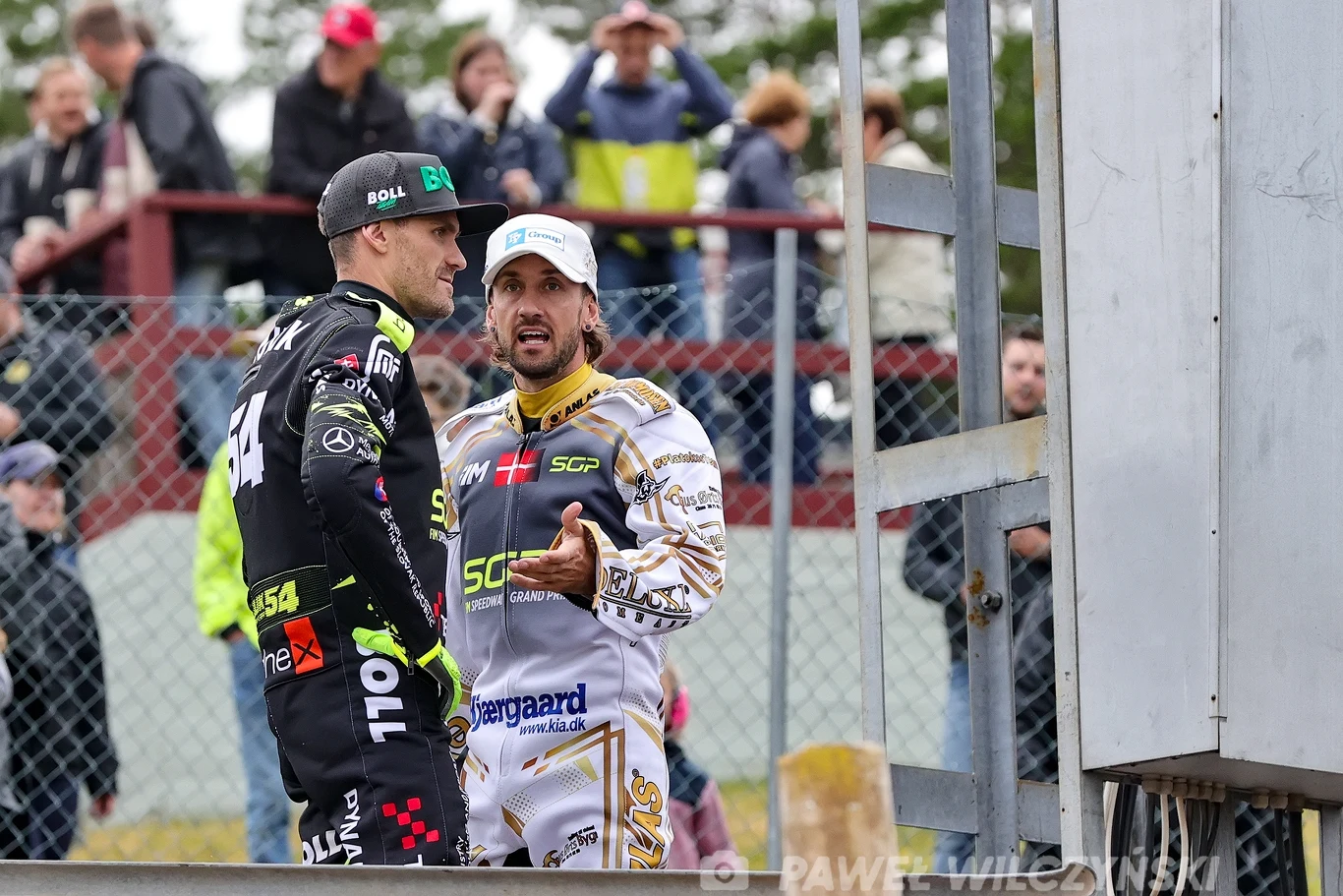 Dwóch żużlowców w kombinezonach z logotypami sponsorów prowadzi rozmowę przy ogrodzeniu toru żużlowego, w tle widoczna publiczność siedząca na trybunach.