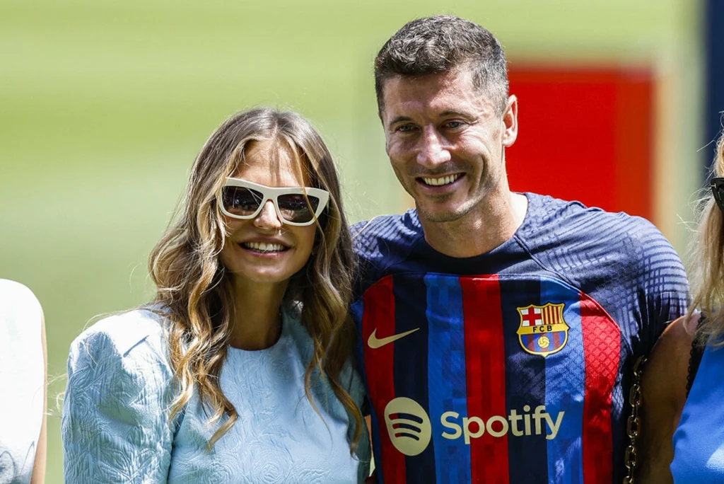 Anna Lewandowska i Robert Lewandowski Uśmiechnięty mężczyzna w koszulce FC Barcelony z logo klubu i sponsora stoi obok kobiety w jasnej sukience i okularach przeciwsłonecznych, oboje pozują do zdjęcia na tle zielonego boiska.