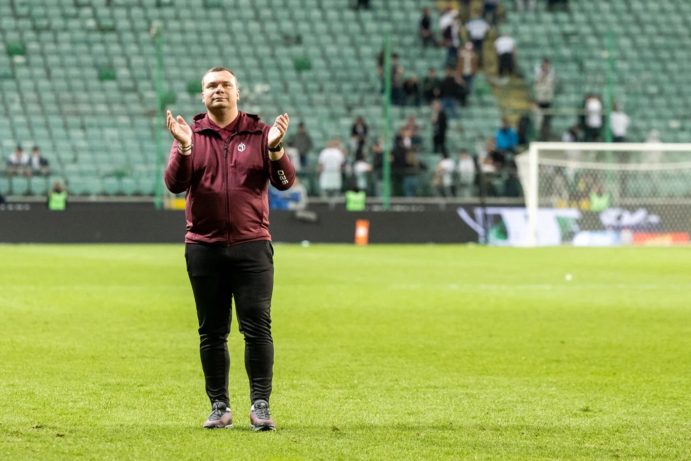 Mężczyzna w bordowej bluzie stoi na murawie stadionu piłkarskiego i bije brawo, w tle widoczne puste trybuny oraz nieliczni kibice.