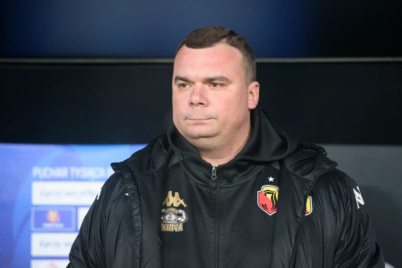 Mężczyzna w kurtce sportowej z logo klubu sportowego stoi na tle rozmazanego wyświetlacza z informacjami.