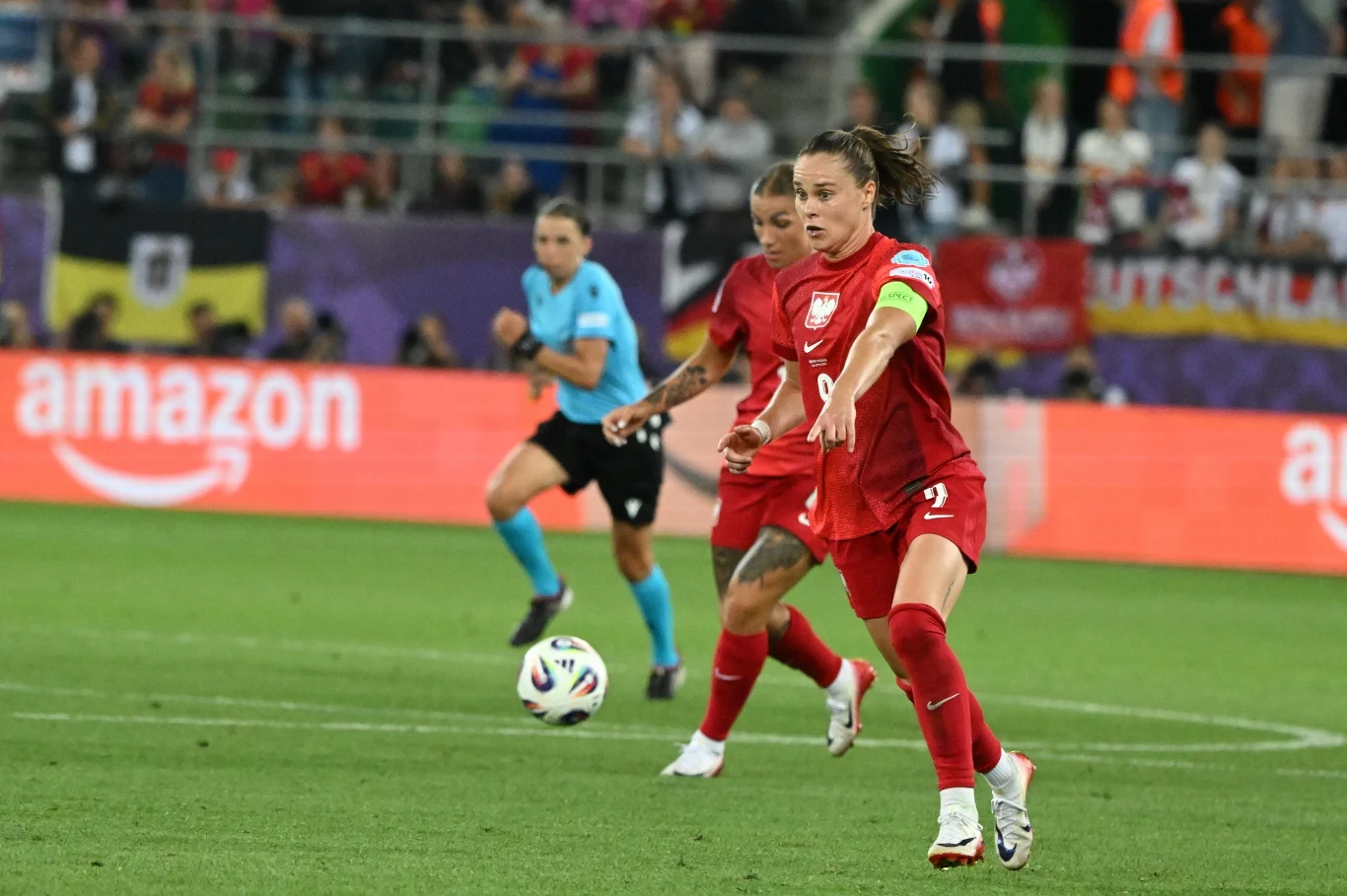 Piłkarka w czerwonym stroju reprezentacji Polski kopie piłkę na murawie podczas meczu piłki nożnej, za nią inna zawodniczka w czerwonym stroju oraz sędzina ubrana na niebiesko, w tle trybuny z kibicami i reklamy.